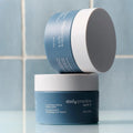 Two blue containers of Daily Practice mattifying balm rest against a tiled background, one stacked on the other. The bottom container highlights nourishing makeup details, crafted as part of the Daily Practice Three Step Eye Care Routine by Dr. B for ophthalmologist-formulated care.