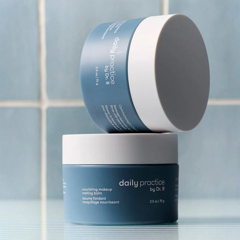 Two blue containers of Daily Practice mattifying balm rest against a tiled background, one stacked on the other. The bottom container highlights nourishing makeup details, crafted as part of the Daily Practice Three Step Eye Care Routine by Dr. B for ophthalmologist-formulated care.