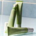 Three tubes of Daily Practice Three Step Eye Care Routine cream are on a striped surface, two upright and one with an open cap showing the applicator. This anti-aging eye cream fits perfectly into your daily routine, set against blue tiles.