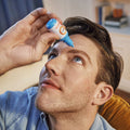 Wearing a blue shirt, a person tilts their head and applies Bausch + Lombs Blink Triple Care 3 in 1 Eye drops from its blue and white bottle into their left eye for lasting hydration. They are indoors with a blurred bookshelf in the background.