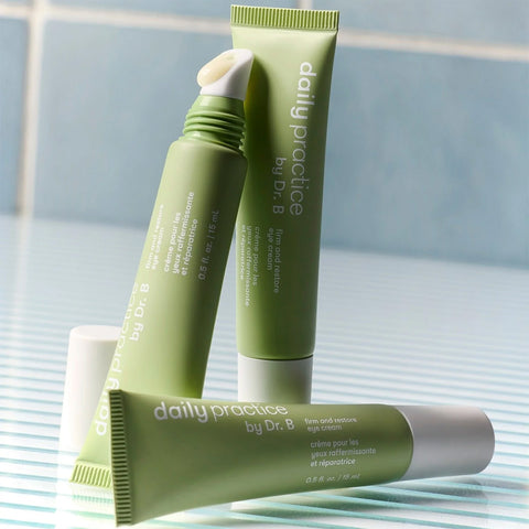 Three green tubes of Daily Practice Firm & Restore Eye Cream are placed on a striped surface. Designed to target fine lines, they have white caps, dispensing tips, and product details in white text against a tiled background.