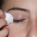 Using LidHygenix Gentle Foam Eyelid Cleanser with a cotton ball, a person softly cleans their closed eyelid, following blue arrows for lid hygiene. The skin looks smooth and the eyelashes remain visible, promoting eye health.