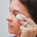 With their eyes closed, a person with long dark hair gently applies the MyboClean Hydrating Lids and Lash Cleansing Gel using a white, oval pad. They wear a silver ring and display a calm expression, while the blurred background adds to the serene atmosphere.