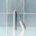 A tube of Daily Practice Lash Enhancing Serum for fuller, longer, healthier lashes (4ml) sits open on a reflective surface with a thin applicator leaning against it. Light blue tiled walls highlight its cutting-edge formula infused with biomimetic peptides for optimal lash growth.