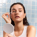 A person with damp hair uses the Daily Practice Lash Enhancing Serum near their eye with a dropper, holding a round mirror while wearing a white tank top against a backdrop of light blue and white tiles, setting a soothing morning scene.