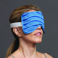 Wearing a Bruder Eye Compress Cold Therapy mask, featuring blue wavy stripes and an elastic band for puffiness reduction, the person has neatly tied-back hair and a sleeveless top against a plain gray background.