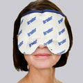 A woman uses the Bruder Moist Heat Sinus Compress over her eyes and nose, set against a plain gray background. The white compress, featuring blue text, is designed with a curved shape for a snug facial fit.
