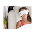 A person in a red top enjoys soothing relief from dry eyes with a Heyedrate Warm Compress Eye Mask by PRN, lying on white bedding with a serene setup of a wooden side table and plant nearby.