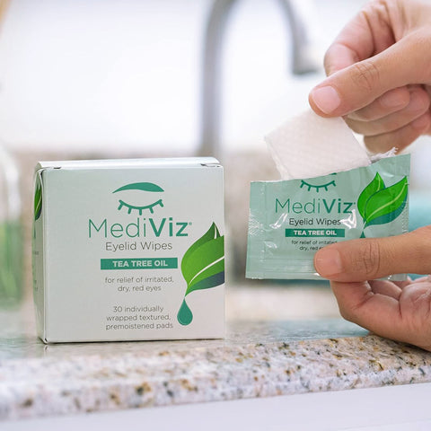 A hand opens a sachet of Mediviz Tea Tree Eyelid Cleansing Wipes by PRN, featuring teal and white packaging designed to relieve irritated, dry eyes. A similar design is visible on a box in the background.