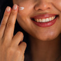 A smiling person applies Merakim Facial Sunscreen SPF 50 to their cheek with fingers, revealing white teeth and red lipstick against a blurred background, while enjoying the protective benefits on sensitive skin.