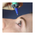Against a dark blue background, a light-skinned person tilts their head back, as a hand holds the Nanodropper Eye Drop Extender Adaptor poised above their eye, reducing drop size by 60% for less waste and easier application.