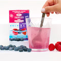A hand stirs a pink Dry Eye Drink in a clear glass, surrounded by fresh blueberries and raspberries. A box labeled Dry Eye Drink™ Complete Set by Bruder with berry imagery suggests the drink is a mixed berry-flavored electrolyte mix, promoting eye health and dry eye relief.