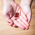 Hands holding EyePromise Macular Health Complete AREDS 2 capsules to enhance vision and support macular health, over a wooden floor background.