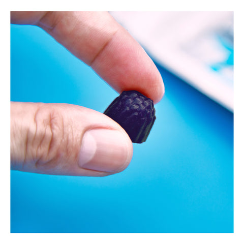 A hand pinches a small, dark candy against a bright blue background, showcasing FitEyez Eye Fitness Gummies. The textured surface suggests the power of Astareal® Astaxanthin for eye health and potential dry eye protection.
