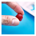 A hand holds a small, wild berry FitEyez gummy against a bright blue background, pinched between the thumb and index finger.