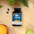 A bottle of Focus Select Soy Free Supplement, AREDS2 formula by Focus Vitamins, rests on wood amid two softgel capsules, an orange, a green apple, and greenery. This supplement is formulated to support macular health.