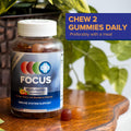 A bottle of Focus Vitamins Multivitamin Gummies rests on a wooden table, its label featuring orange, cherry, and strawberry flavors for immune health and energy. A sign reads, Chew 2 gummies daily with a meal. Green plants provide a refreshing backdrop.