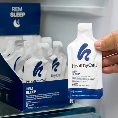 A hand reaches for a HealthyCell REM Sleep Supplement and Sleep Aid gel pack from a box in the refrigerator. The white packet with blue branding contains calming herbs, amino acids, and minerals to support sleep cycles, all with a delightful cherry flavor.