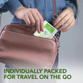 A Noveha Tea Tree Oil Eyelid & Lash Wipe packet is slipped into a pocket of a light pink handbag. The text at the bottom reads, Individually packed with natural ingredients for travel on the go.