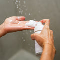 Close-up of a person dispensing Merakim Tea Tree Oil Foam Facial Cleanser (120mL) onto their palm, with water droplets in the background, suggesting a wet shower or washing setting.
