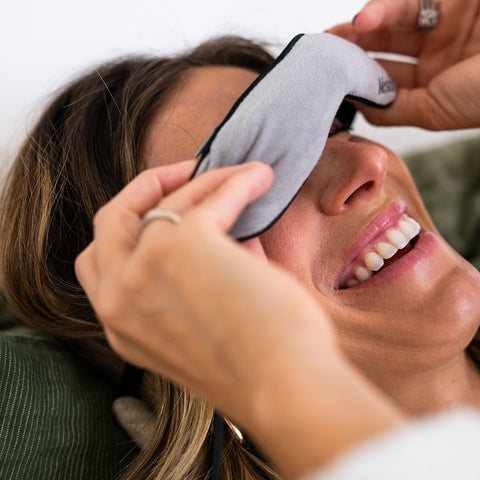A woman smiles while lying down and adjusting the Merakim Professional USB Heat Eye Mask, an adjustable gray heat compress for dry eyes. Her engagement ring glistens on her left hand, enhancing the serene setting.