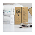 A We Love Eyes Calm Eyelid & Eyelash Cleansing Spray with HypoChlorous (50mL) bottle and box are on a white surface, showcasing its vegan, cruelty-free formula. Chamomile adds soothing properties, while blurred glasses and towels in the background evoke everyday elegance.