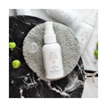 A We Love Eyes Calm Eyelid & Eyelash Cleansing Spray with HypoChlorous (50mL) bottle sits on a gray circular cloth pad on a marble surface, surrounded by three small green fruits and an open cap, with more bottles in the background.