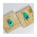 Two turquoise We Love Eyes Focused Eye Roll products with heart-shaped cutouts rest on square marble tiles. Their metallic tops, mimicking stainless steel, are set at a diagonal angle on a white background, enhancing elegance.
