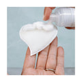 Someone dispenses foam onto a We Love Eyes Eyelid Scrub Mitt, which is heart-shaped and textured, resembling the gentle care of an eyelid scrub. The scene is set against a soft pastel background.