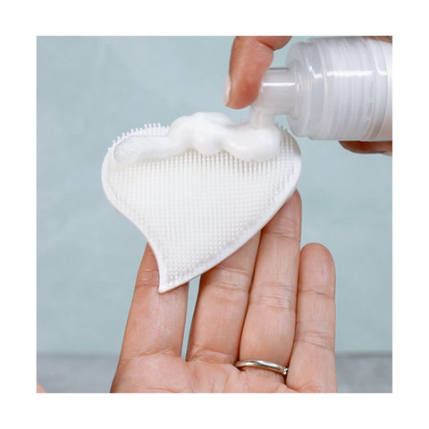 Someone dispenses foam onto a We Love Eyes Eyelid Scrub Mitt, which is heart-shaped and textured, resembling the gentle care of an eyelid scrub. The scene is set against a soft pastel background.