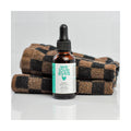 A small brown dropper bottle labeled We Love Eyes is placed in front of folded checkered towels. It contains We Love Eyes 100% All Natural Tea Tree Makeup Remover Oil with organic jojoba oil, perfect for eyelid cleaning and removing waterproof makeup, in a 30mL capacity.