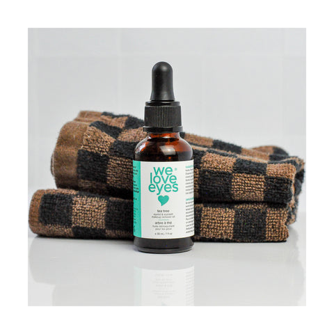A small brown dropper bottle labeled We Love Eyes is placed in front of folded checkered towels. It contains We Love Eyes 100% All Natural Tea Tree Makeup Remover Oil with organic jojoba oil, perfect for eyelid cleaning and removing waterproof makeup, in a 30mL capacity.
