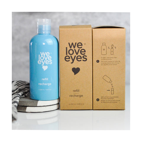 A blue We Love Eyes bottle, labeled Tea Tree Eyelid Foaming Cleanser Refill (250mL), is accompanied by two identical brown boxes. The box on the right displays easy-to-follow instructions. A striped cloth and stacked coasters in the background enhance the elegant setup.